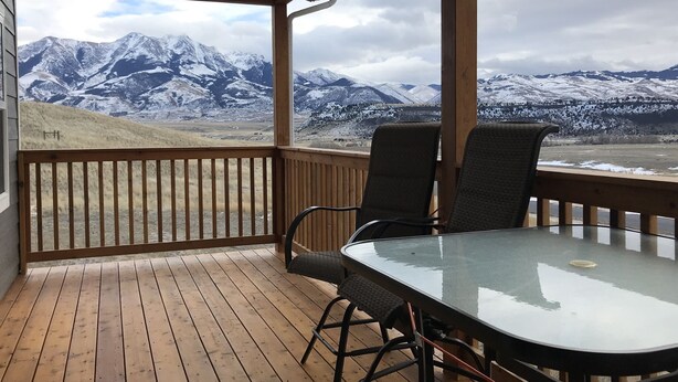 A wooden deck with a glass table and chairs, overlooking a snowy mountain range.