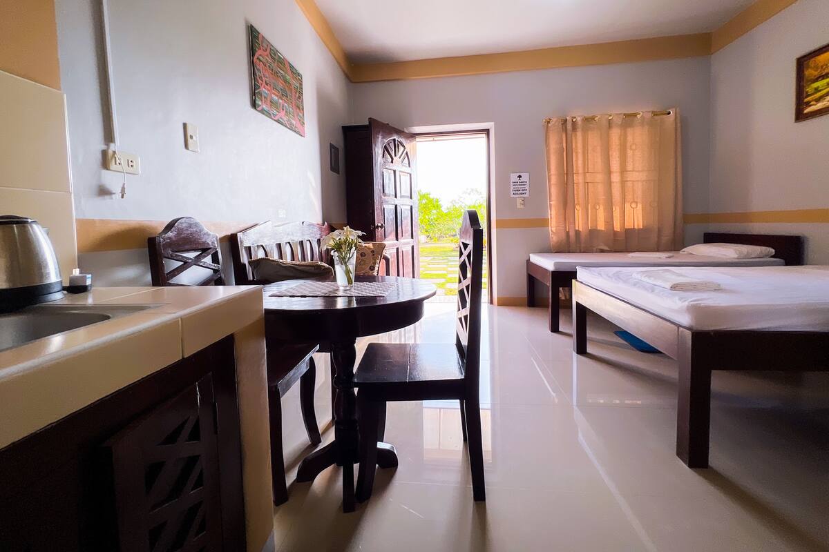 A hotel room with a dining table, two beds, a kitchen area, and a view of the outdoors.