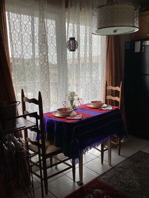 Un coin repas avec une table dressée pour deux personnes, un lustre, des rideaux en dentelle et un réfrigérateur.