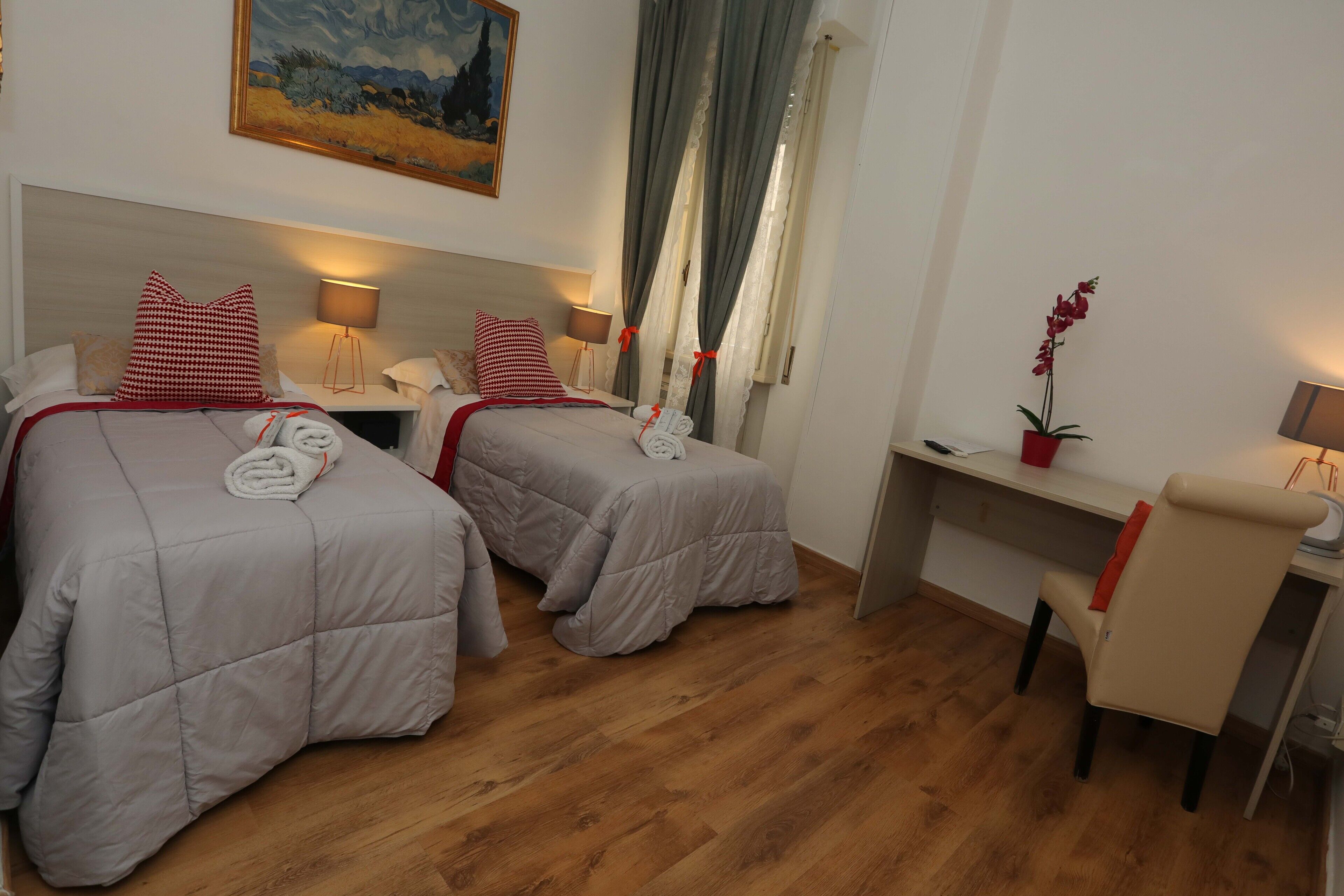 A hotel room with two beds, a desk, a chair, a lamp, a potted plant, and a painting on the wall.