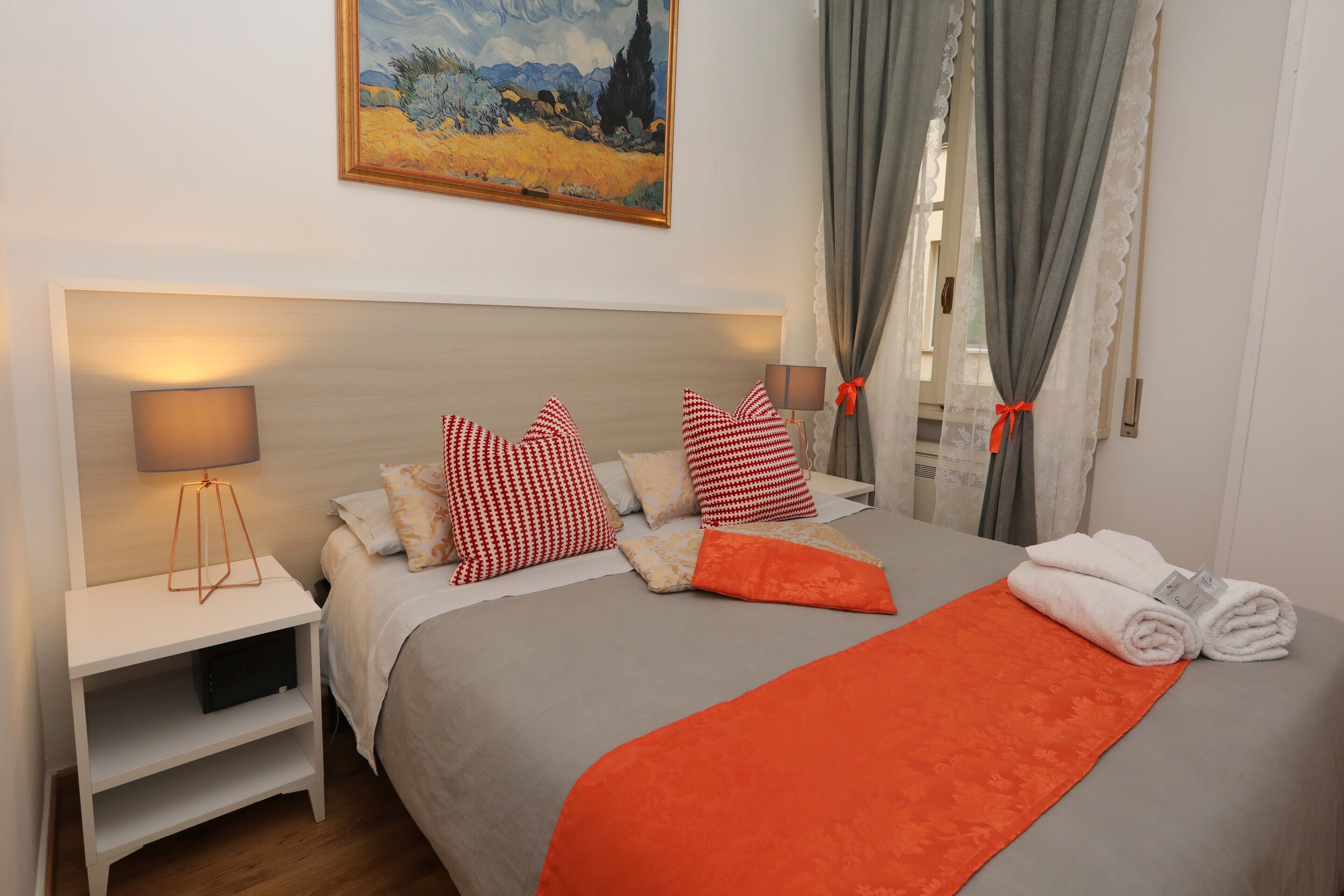A neatly made bed with red and white checkered pillows, a grey and orange blanket, and a pair of folded towels on the side.