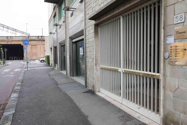 A street view with a closed metal gate, a bicycle lane, and a building with a sign reading "Dimora".
