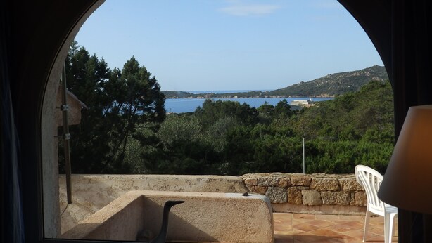 A view through a circular window of a stone patio with a white chair, overlooking a coastal landscape with trees and a clear sky. - Villa near the sea, beautiful view, two terraces (PIANOTTOLI CALDARELLO)