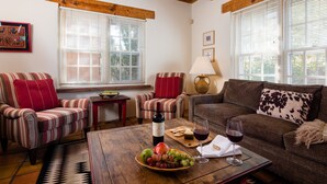 A cozy living room with striped sofas, a wooden coffee table with fruit and wine, and large windows with blinds. - Light Filled, Spacious Casita~Walk to Downtown & Mountain Views (Santa Fe)