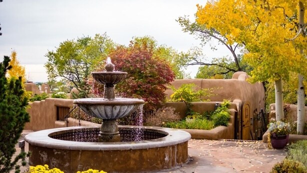A circular stone fountain with water spouts, surrounded by a stone patio and yellow flowers. - Light Filled, Spacious Casita~Walk to Downtown & Mountain Views (Santa Fe)