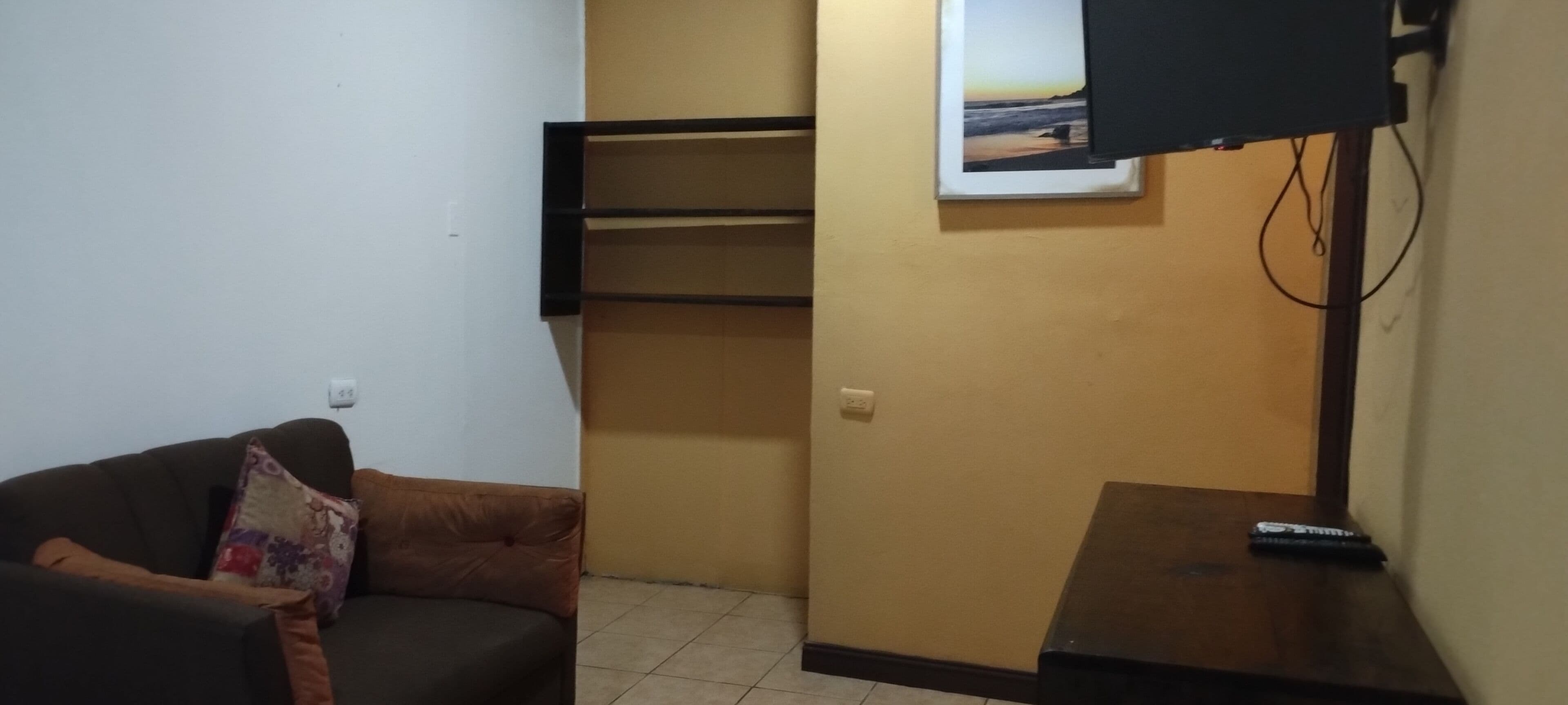 Una pequeña sala de estar con un sofá, un televisor montado en la pared y un control remoto sobre una mesita.