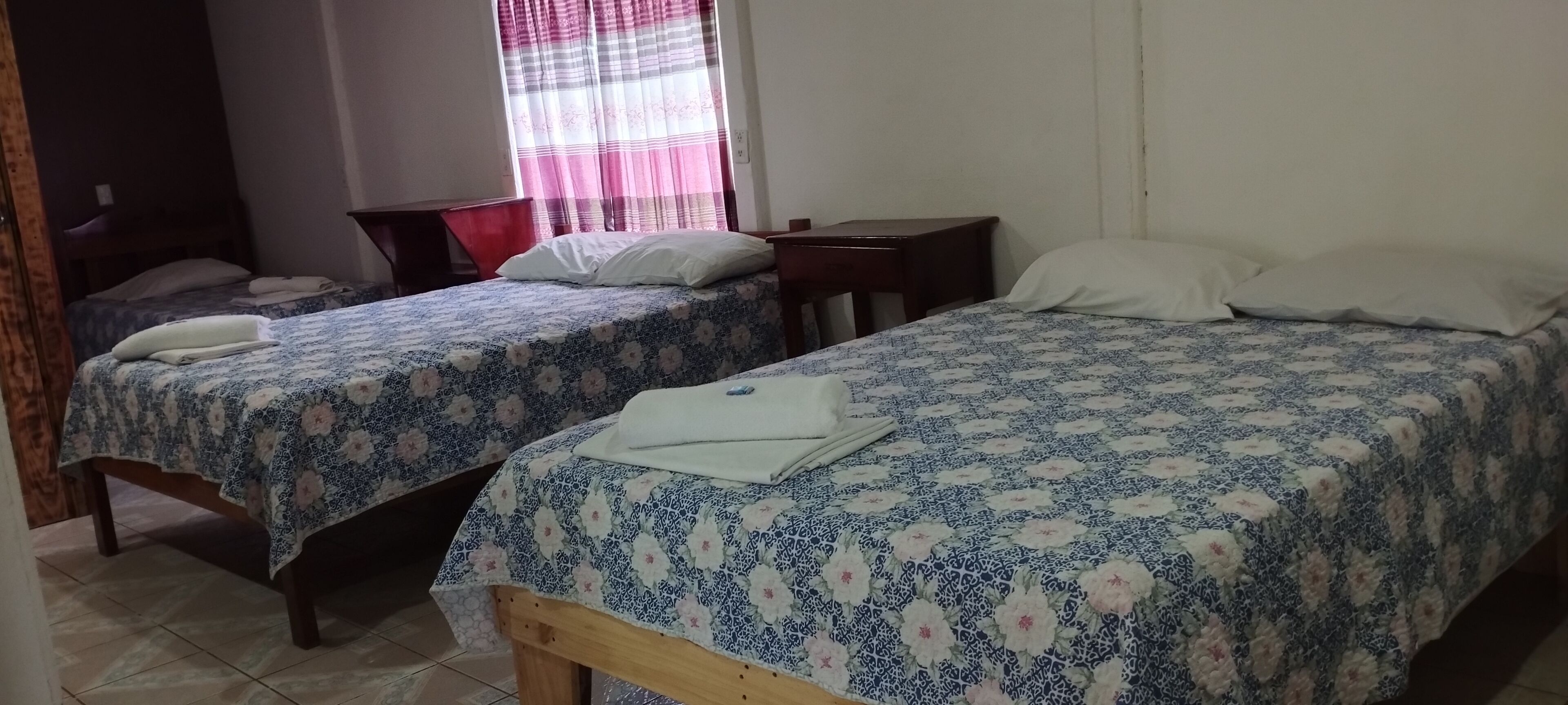 A room with two beds, each with a floral-patterned bedspread, a wooden bench, a desk, and a chair.