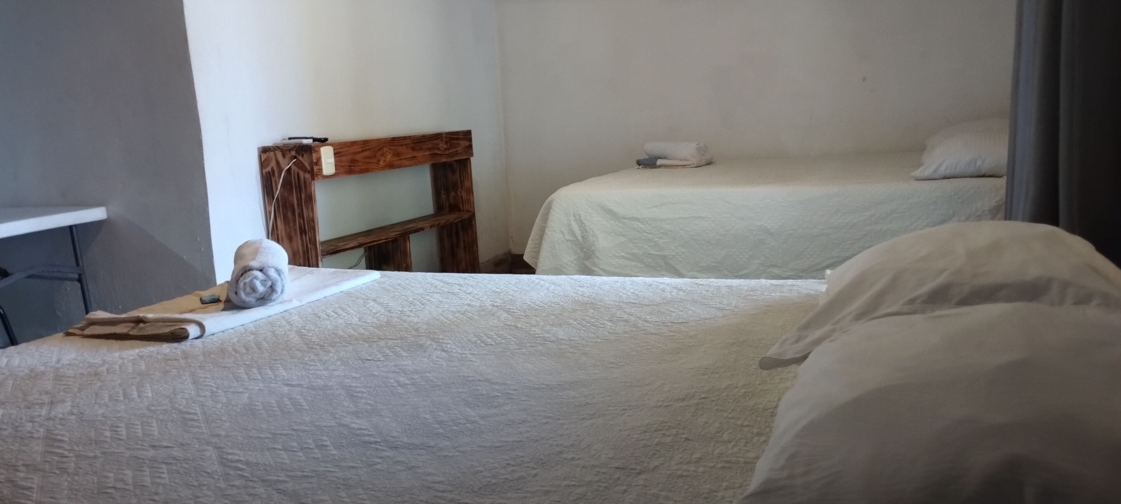 Una habitación con dos camas, cabecero de madera, un escritorio con una toalla y un libro, y un estante pequeño.