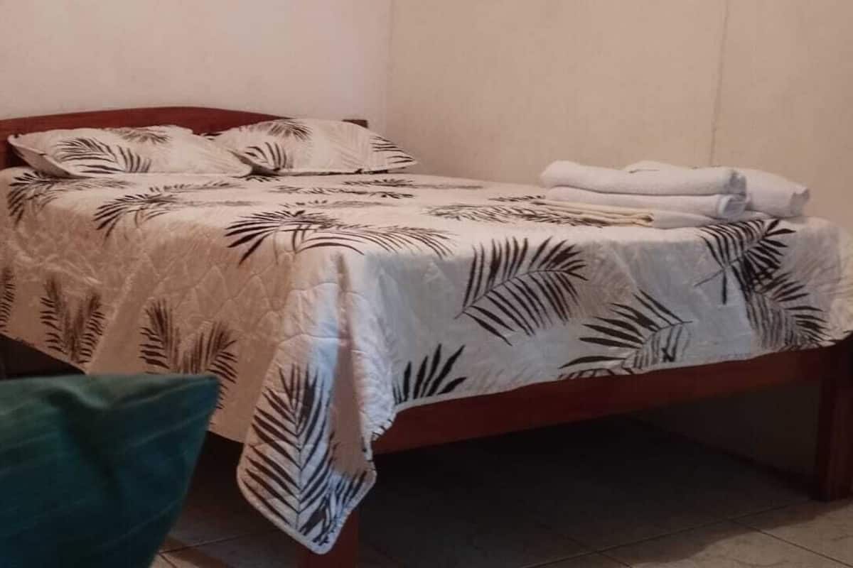 A single bed with a leaf-patterned bedspread, a green pillow, and a stack of white towels on the side.