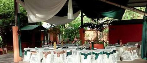 An outdoor covered area with tables set up for an event, surrounded by greenery.