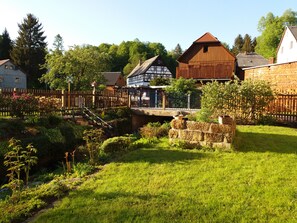 Un village européen traditionnel, avec un pont en bois, des murs de pierre et une végétation luxuriante.