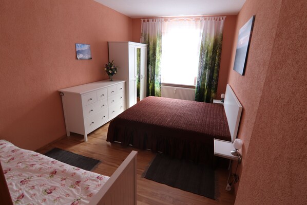 Une chambre à coucher comprenant un lit, une commode, une table de chevet, un radiateur et une fenêtre avec des rideaux.
