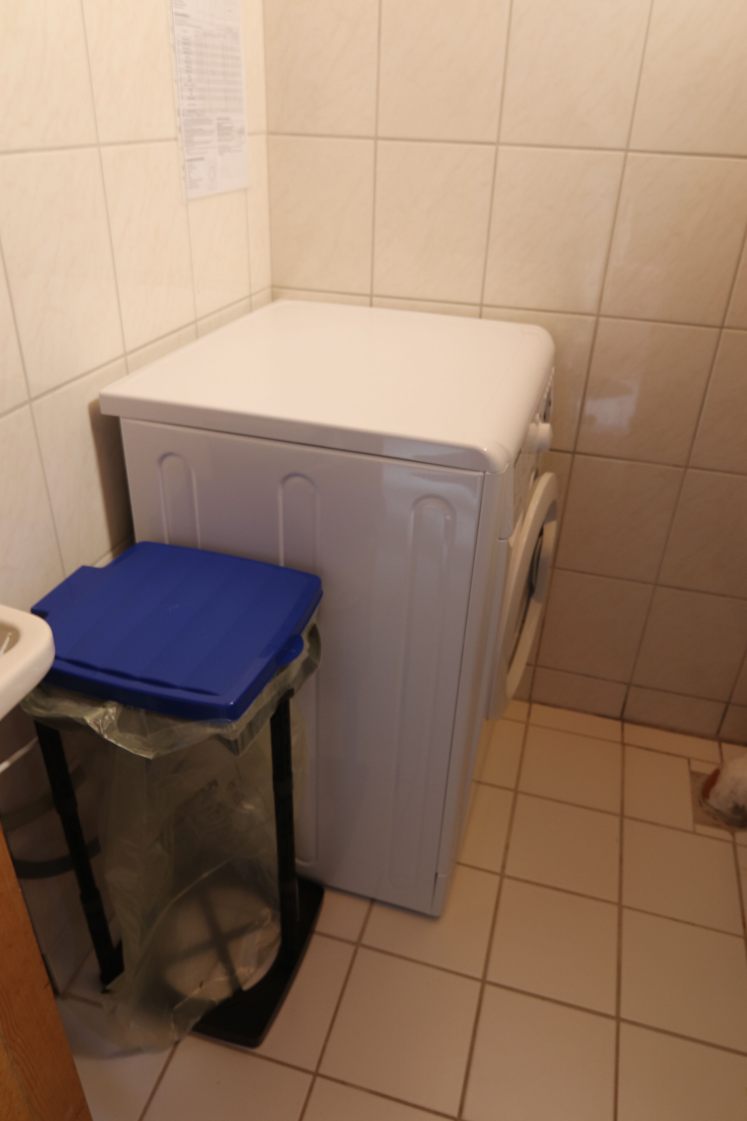 A compact laundry area with a washing machine, a blue drying rack, and a trash bin.