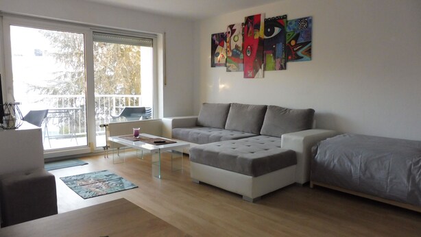 Ein modernes Wohnzimmer mit einem grauen Ecksofa, einem Glastisch und einem Balkon mit Sitzgelegenheiten.