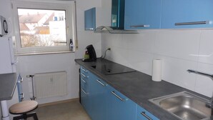 Eine Küche mit blauen Schränken, einer Kaffeemaschine, einem Wasserkocher, einer Spüle und einem Fenster mit Blick auf Gebäude.