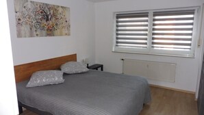 Ein Schlafzimmer mit Bett, Kissen, Nachttisch, Lampe, Heizkörper und einem Fenster mit Jalousien.