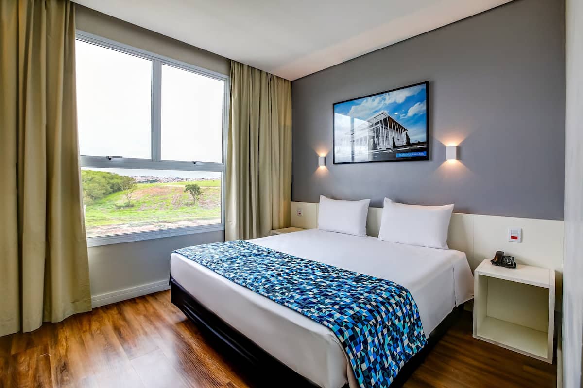 A hotel room with a large bed, a window with curtains, a wall-mounted picture, and a bedside table with a phone.