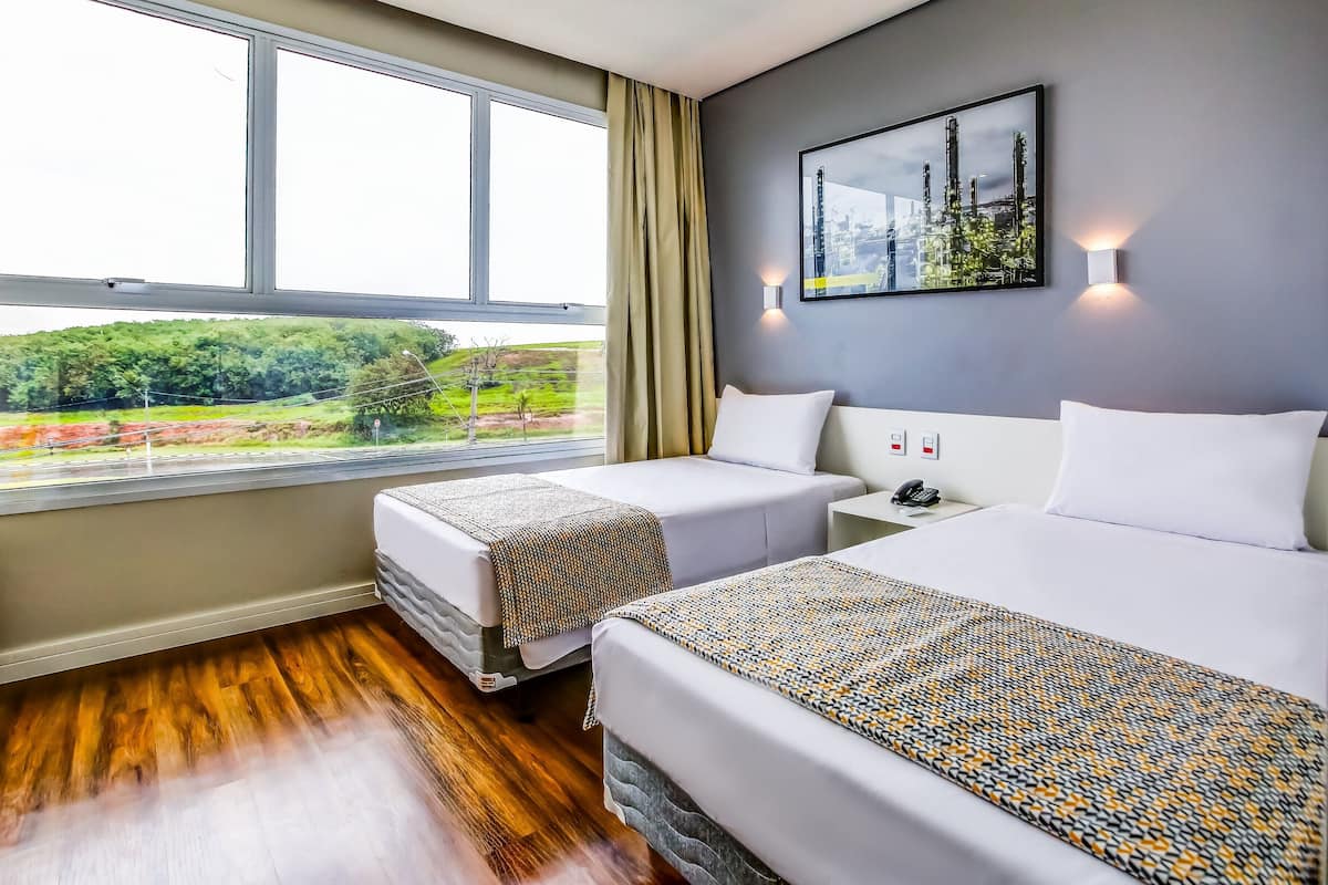 A hotel room with two beds, a large window with a view of greenery, a wall-mounted picture, and a bedside table with a phone.
