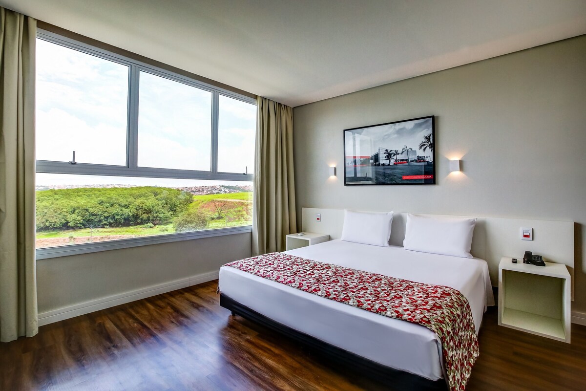 Una habitación de hotel con un ventanal grande, una cama con colcha estampada, una mesita de noche y una lámpara de pared.