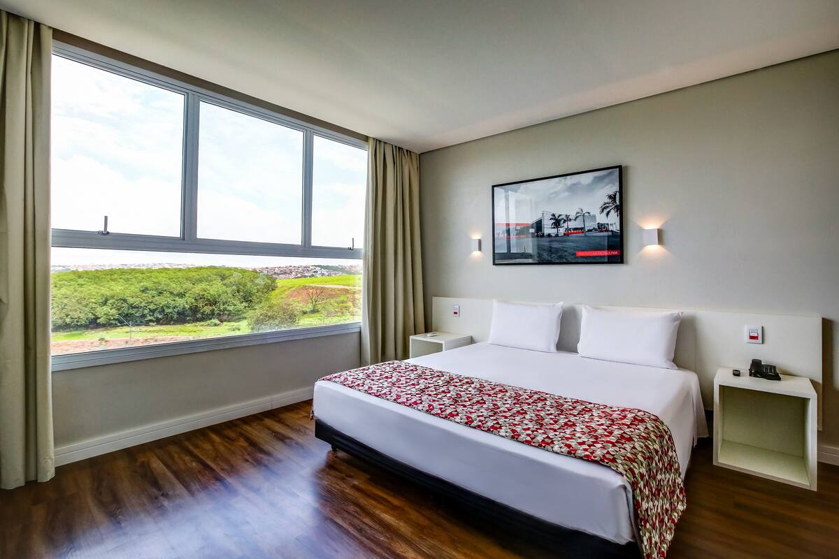 A hotel room with a large window, a bed with a floral bedspread, a nightstand, and a wall-mounted light.