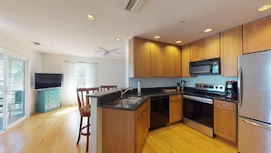 A modern kitchen with wooden cabinets, a black countertop, stainless steel appliances, and a dining area with a TV and teal dresser. - AMAZING RATES & RAVE REVIEWS! Stunning private pool! 3min walk to the Beach 🏖 (Fort Myers Beach)