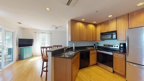 Una cocina moderna con armarios de madera, encimera negra, electrodomésticos de acero inoxidable y un comedor con televisor y cómoda azul aguamarina.