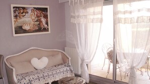 Una habitación con sofá, una pequeña mesa redonda, una silla y un ventanal con cortinas traslúcidas.