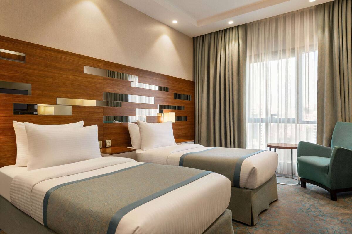 Habitación de hotel con dos camas, cabecero de madera, una mesita pequeña y un sillón verde.