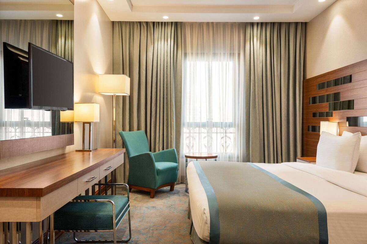 Habitación de hotel con cama, escritorio, silla, televisión y ventana con cortinas.