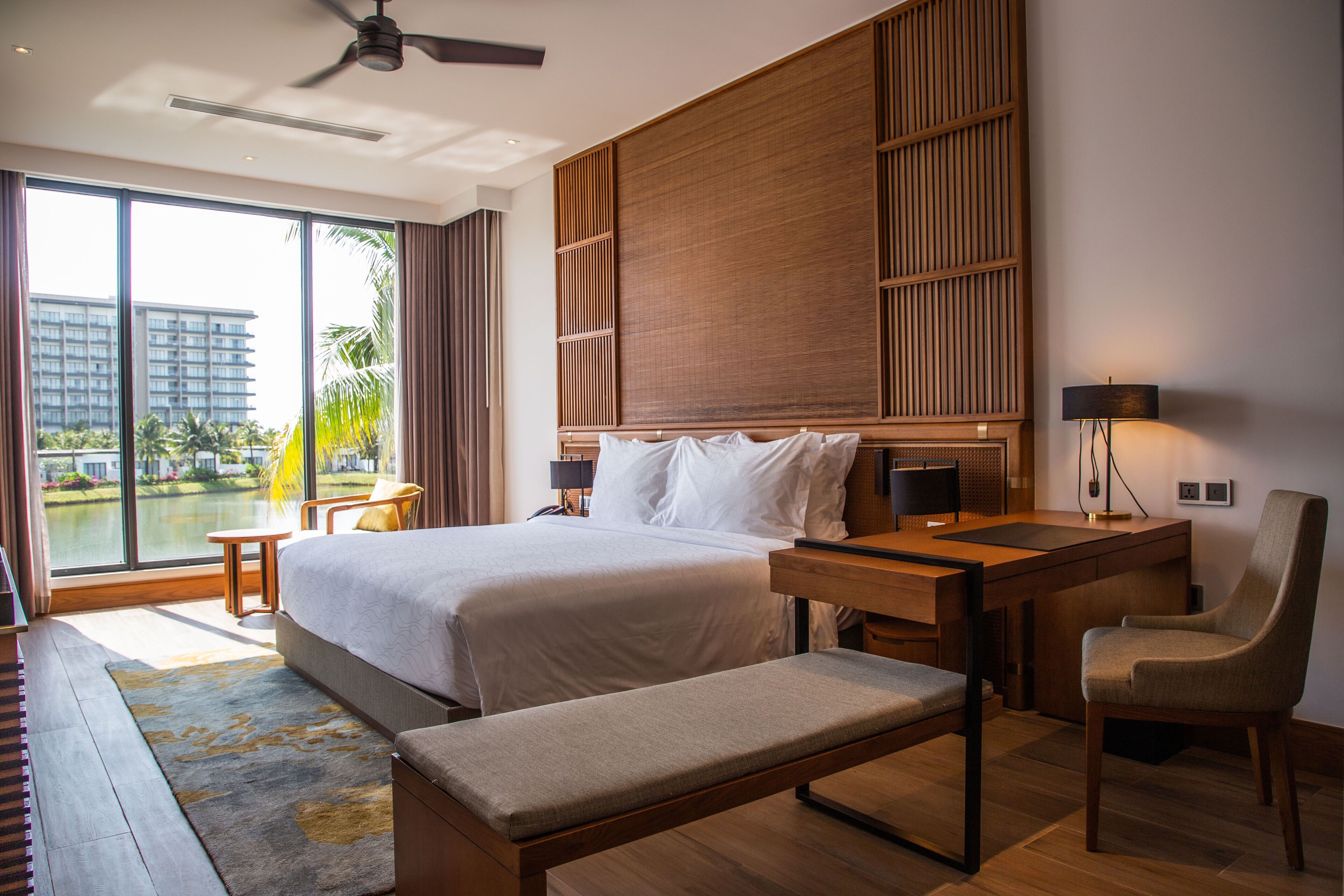 Ein modernes Hotelzimmer mit einem großen Bett, einem Schreibtisch, einem Stuhl, einer Bank und Blick auf ein Gewässer sowie Gebäude.