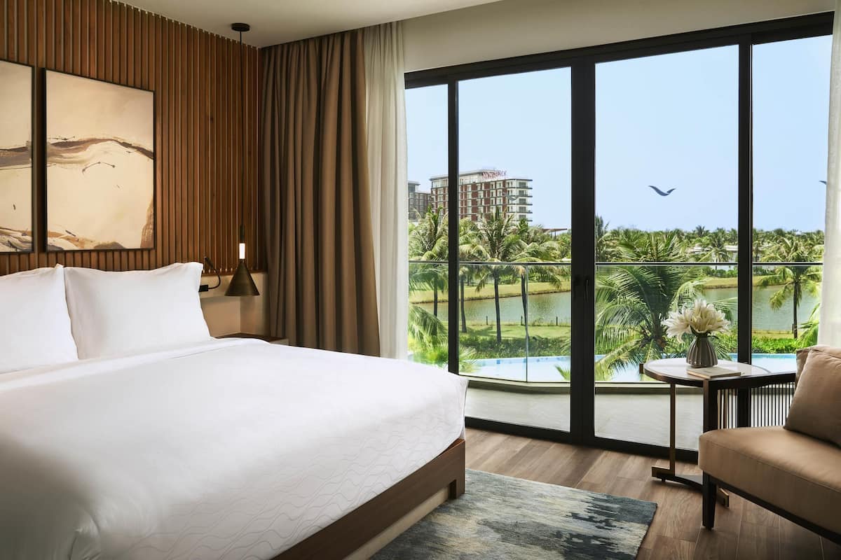 A hotel room with a large bed, a chair, a small table, a painting, and a view of a building and greenery.