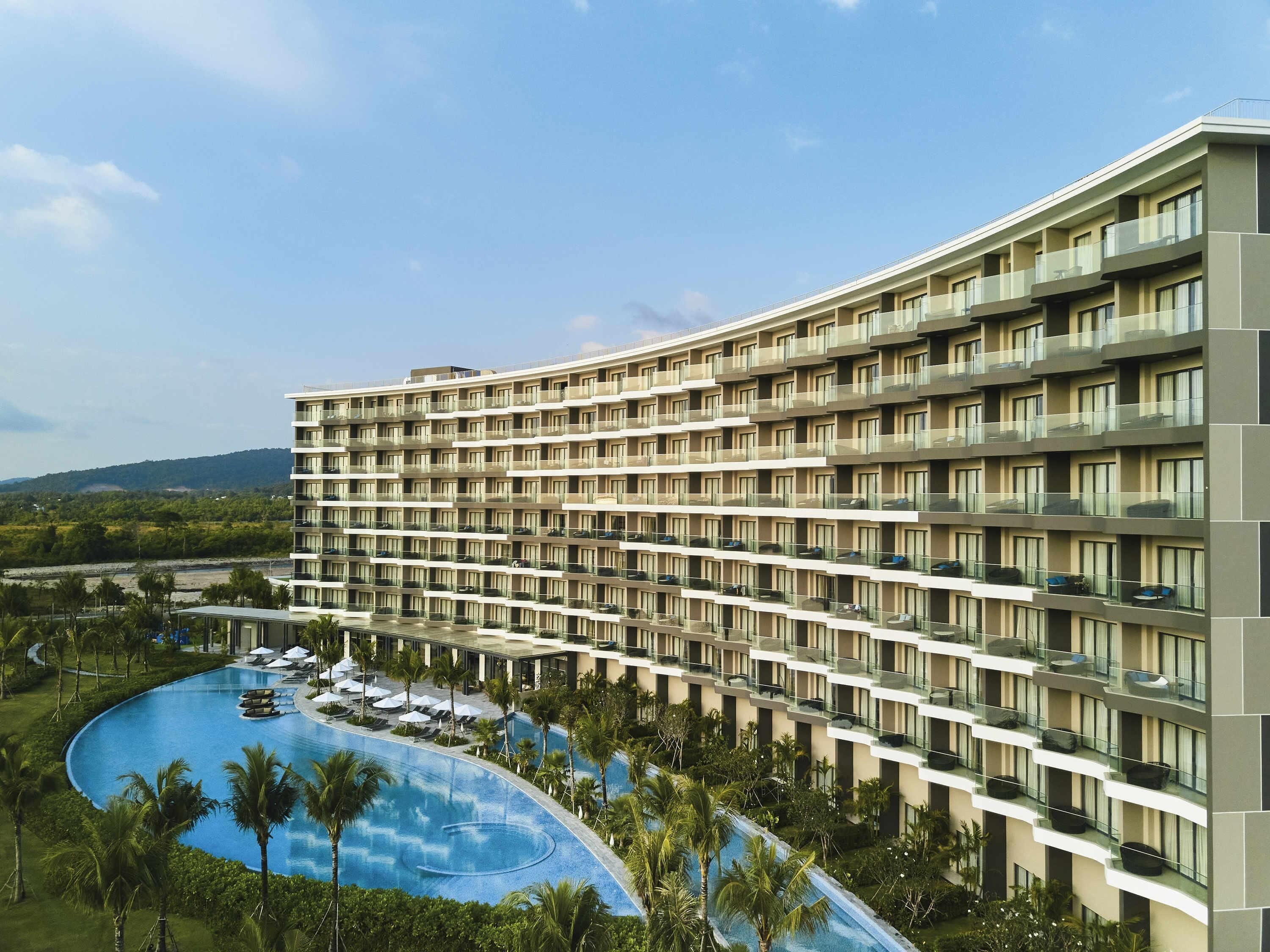 Un hotel con varios pisos, piscina y un área ajardinada con palmeras.