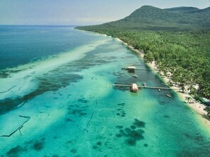 Vue aérienne d’une plage tropicale aux eaux turquoise et limpides, bordée d’un rivage sablonneux, avec une colline boisée en arrière-plan.