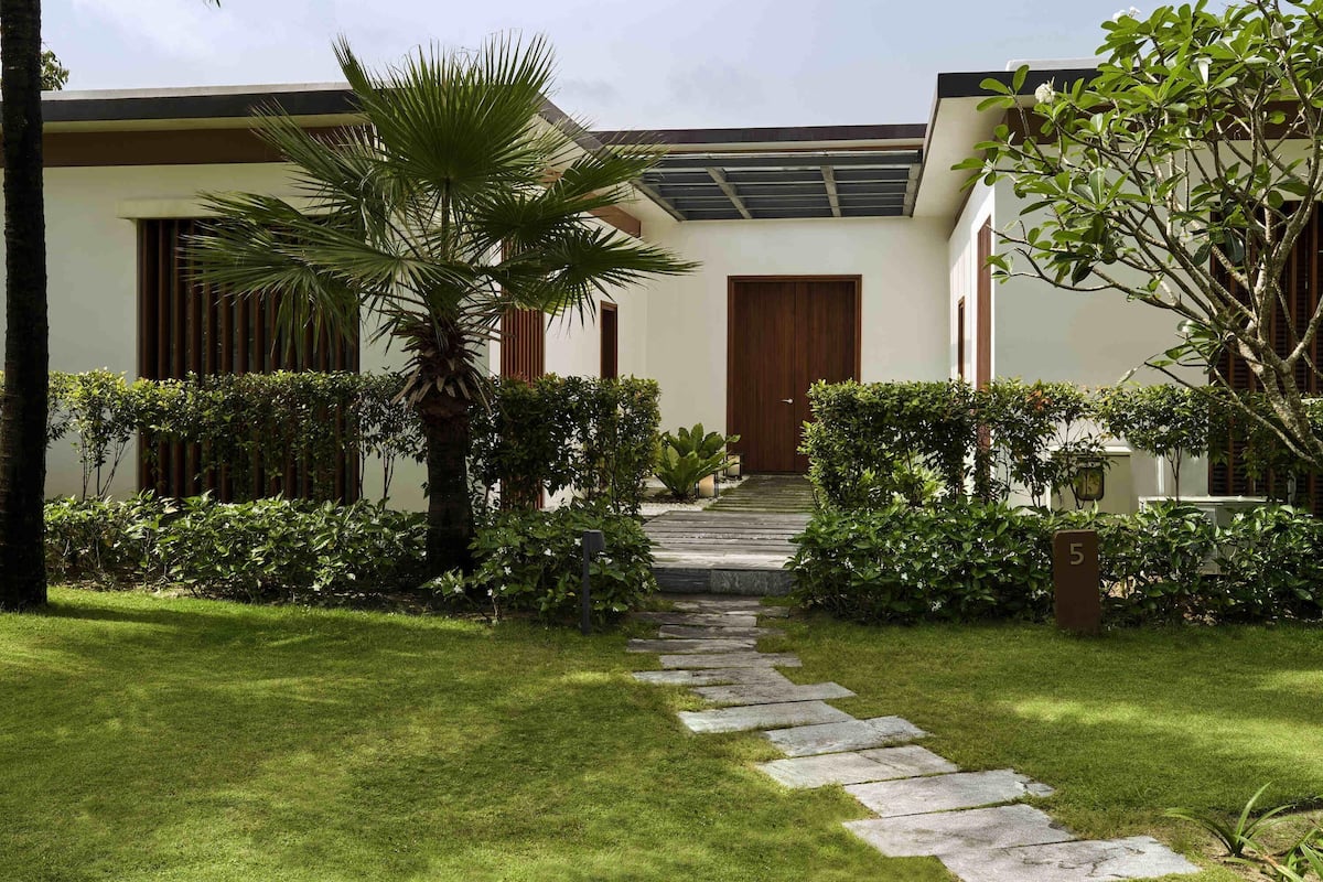A modern building with a stone pathway leading to a wooden door, surrounded by lush greenery and palm trees.