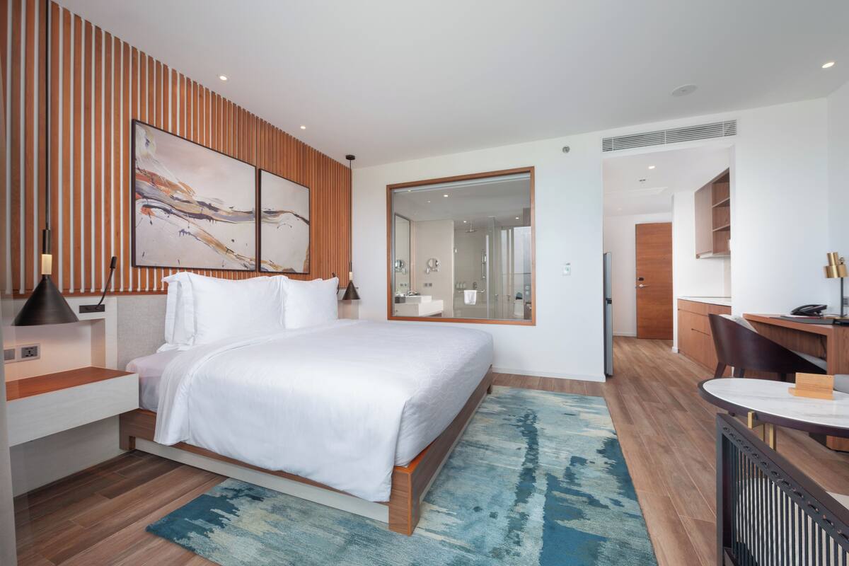 Ein modernes Hotelzimmer mit einem großen Bett, einem Schreibtisch und einem durch eine Glastür einsehbaren Badezimmer.