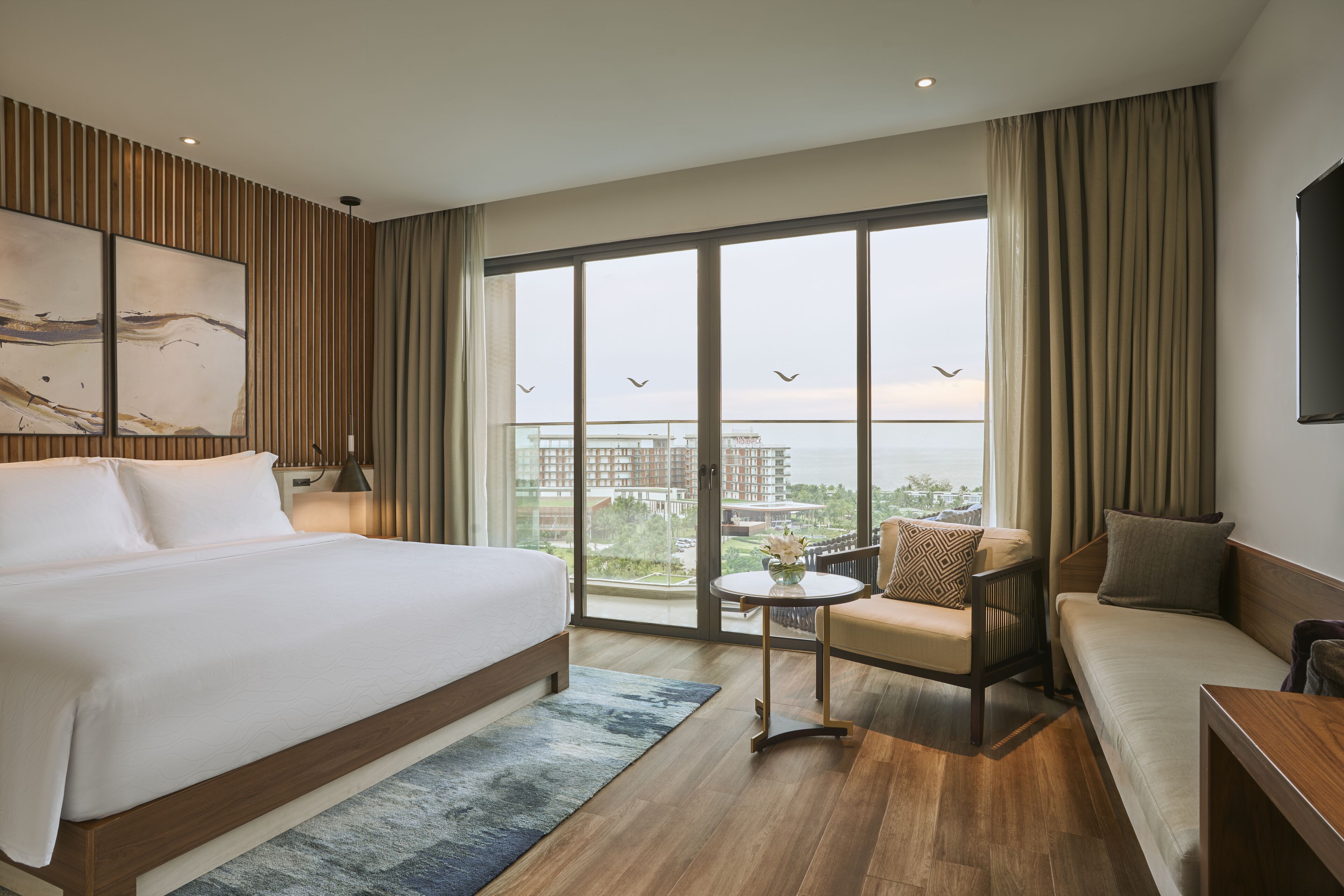 A modern hotel room with a large bed, a sofa, a small table, and a balcony with a view of the city.