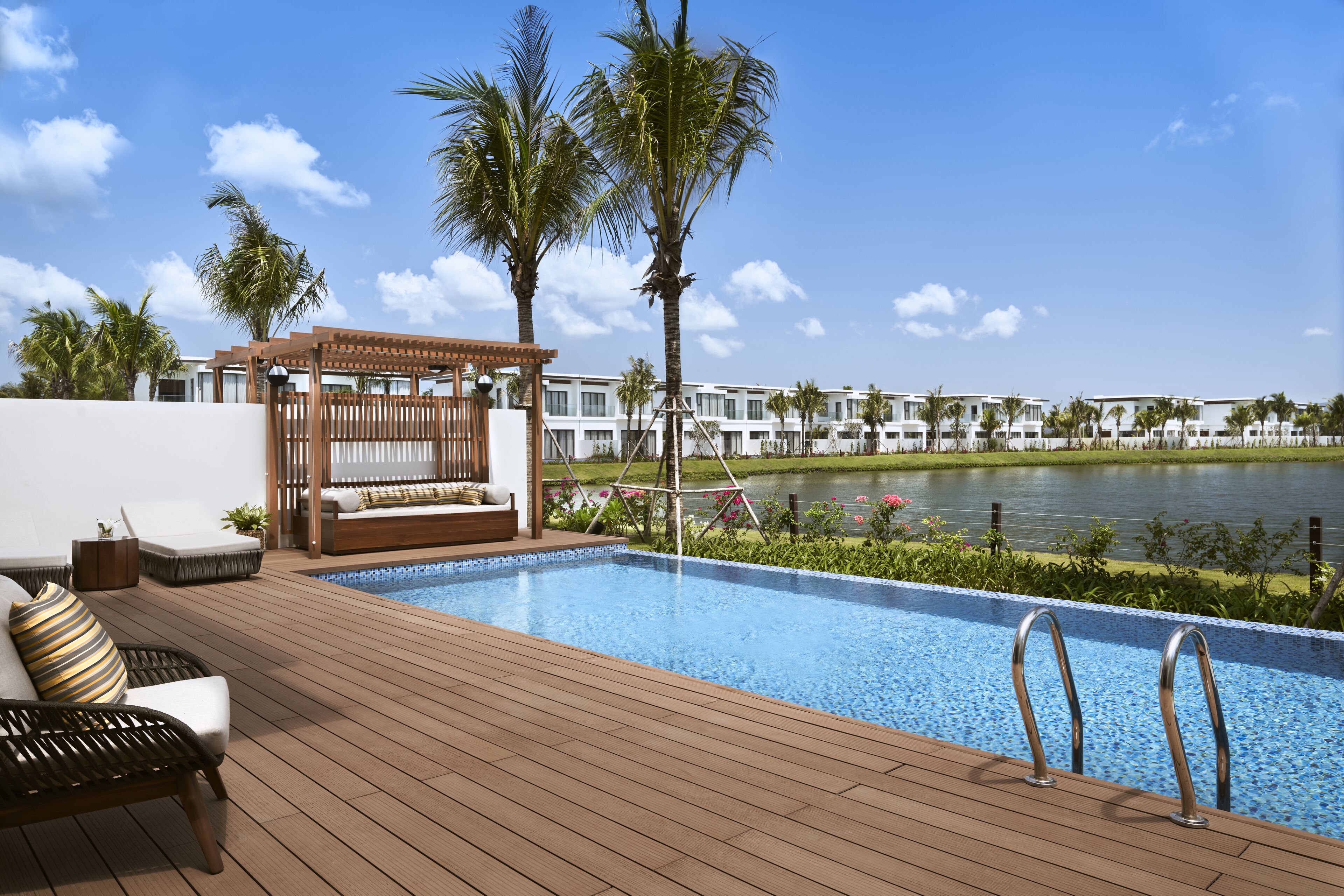 Zona de piscina con terraza de madera, sillas de descanso, un pérgola y vistas a una masa de agua y edificios de hotel.