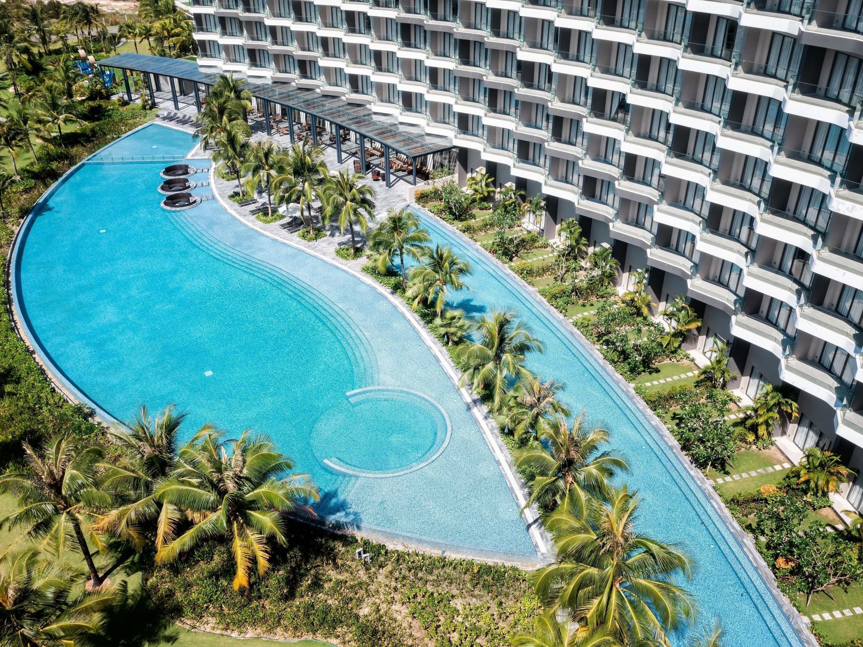 Vista aérea de un hotel con una amplia piscina curva rodeada de palmeras y un edificio de varios pisos con balcones.