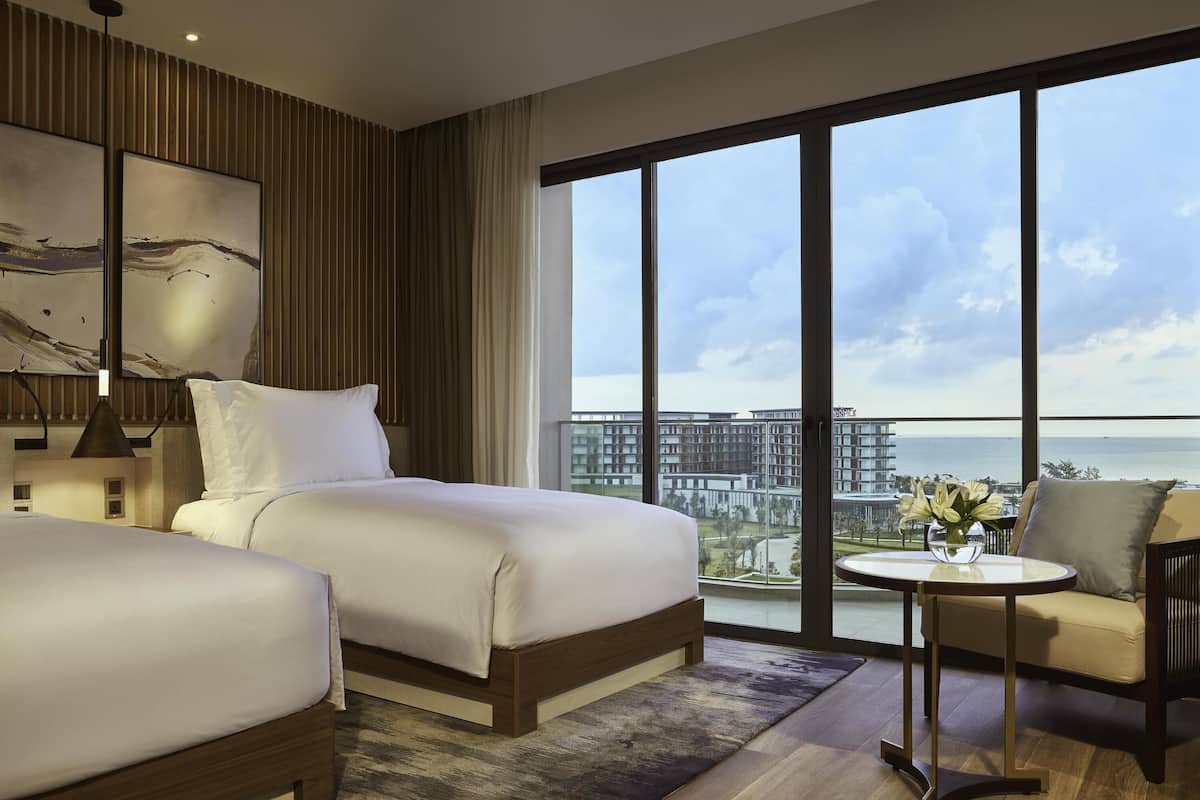 A hotel room with two beds, a small table with a vase, and a large window with a view of buildings and water.