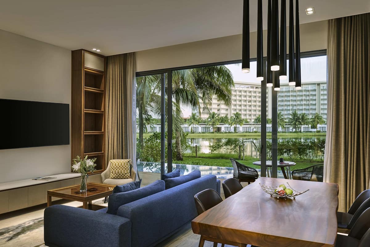 A modern living room with a large TV, a sofa, a dining table, and a view of a hotel building and palm trees.