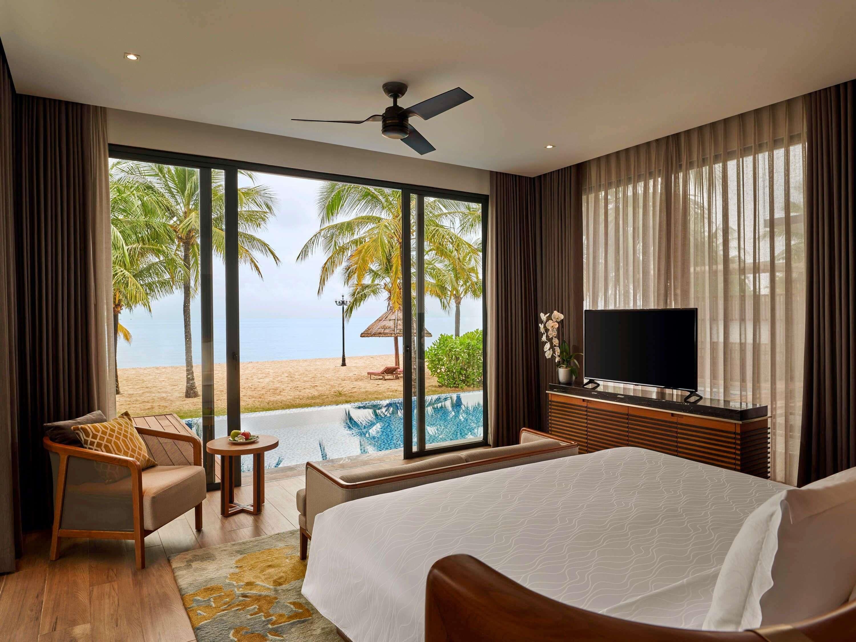 A hotel room with a bed, a TV, a small table, a chair, and a view of a beach and pool.
