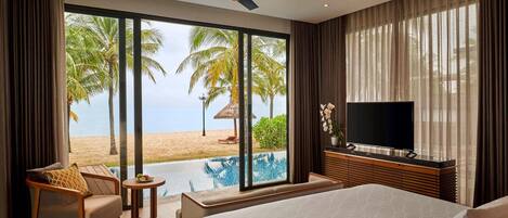 A hotel room with a bed, a TV, a small table, a chair, and a view of a beach and pool.