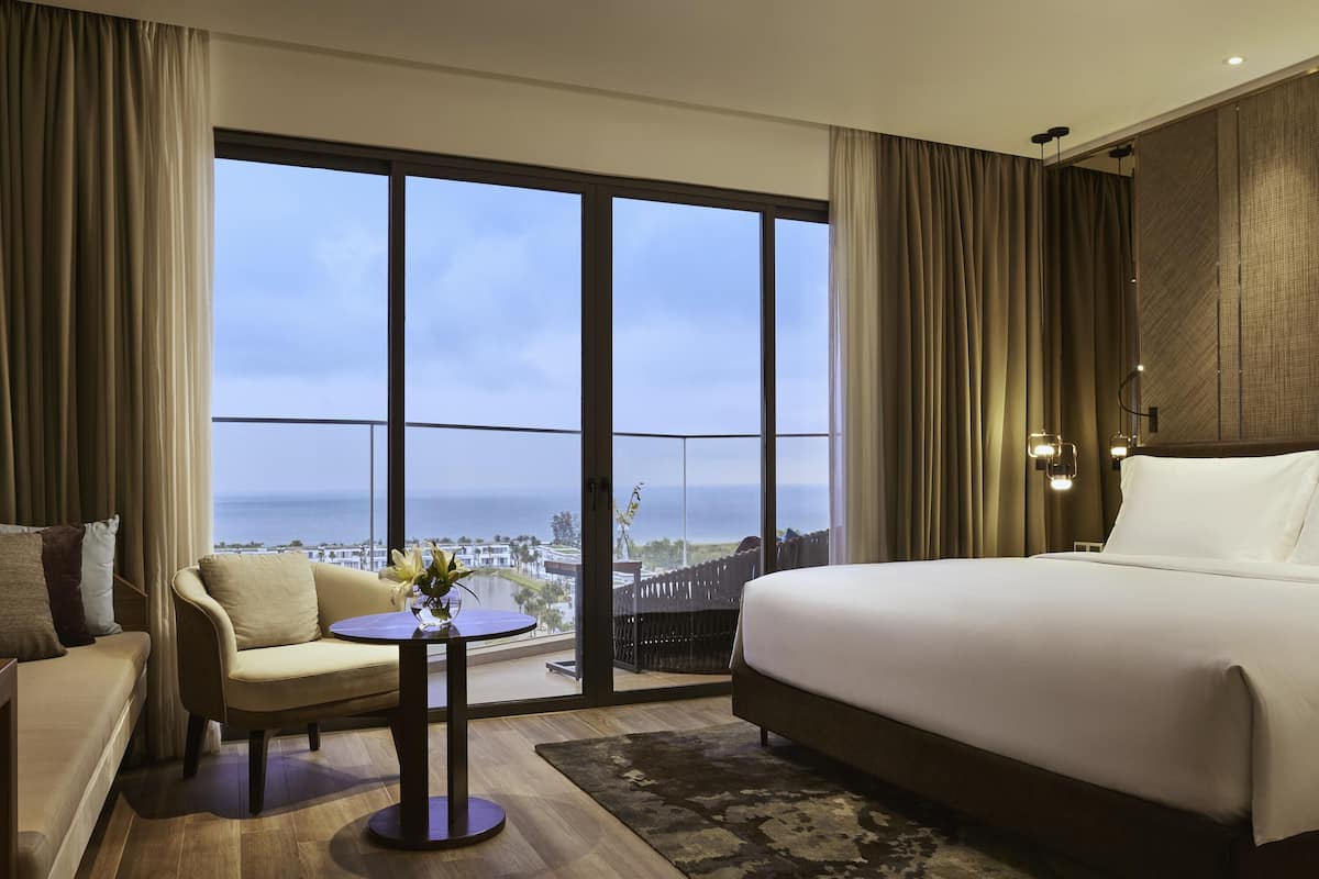 A hotel room with a large bed, a sofa, a small table with a vase, and a view of the ocean.