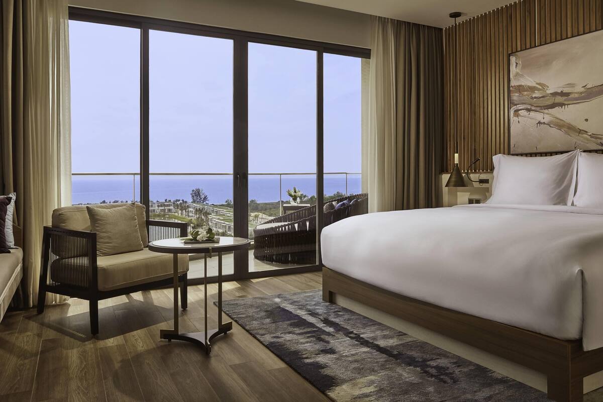 A hotel room with a large bed, a small table, a chair, and a view of the ocean.