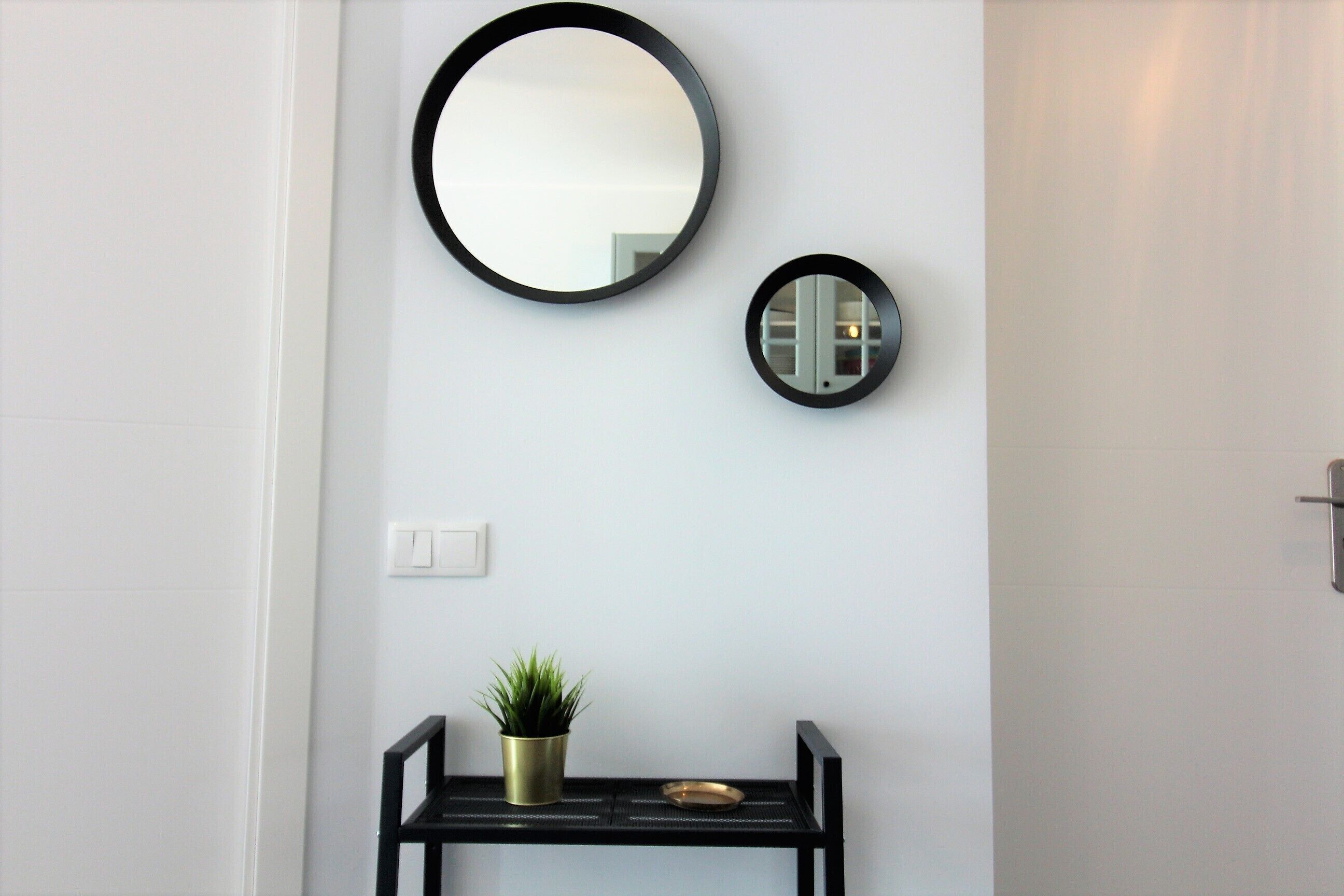 A wall-mounted mirror, a small round mirror, and a potted plant on a black console table.