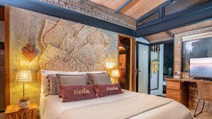Un dormitorio con un amplio cabecero tejido, dos lámparas de noche, una cama con varias almohadas y un escritorio de madera con una silla.