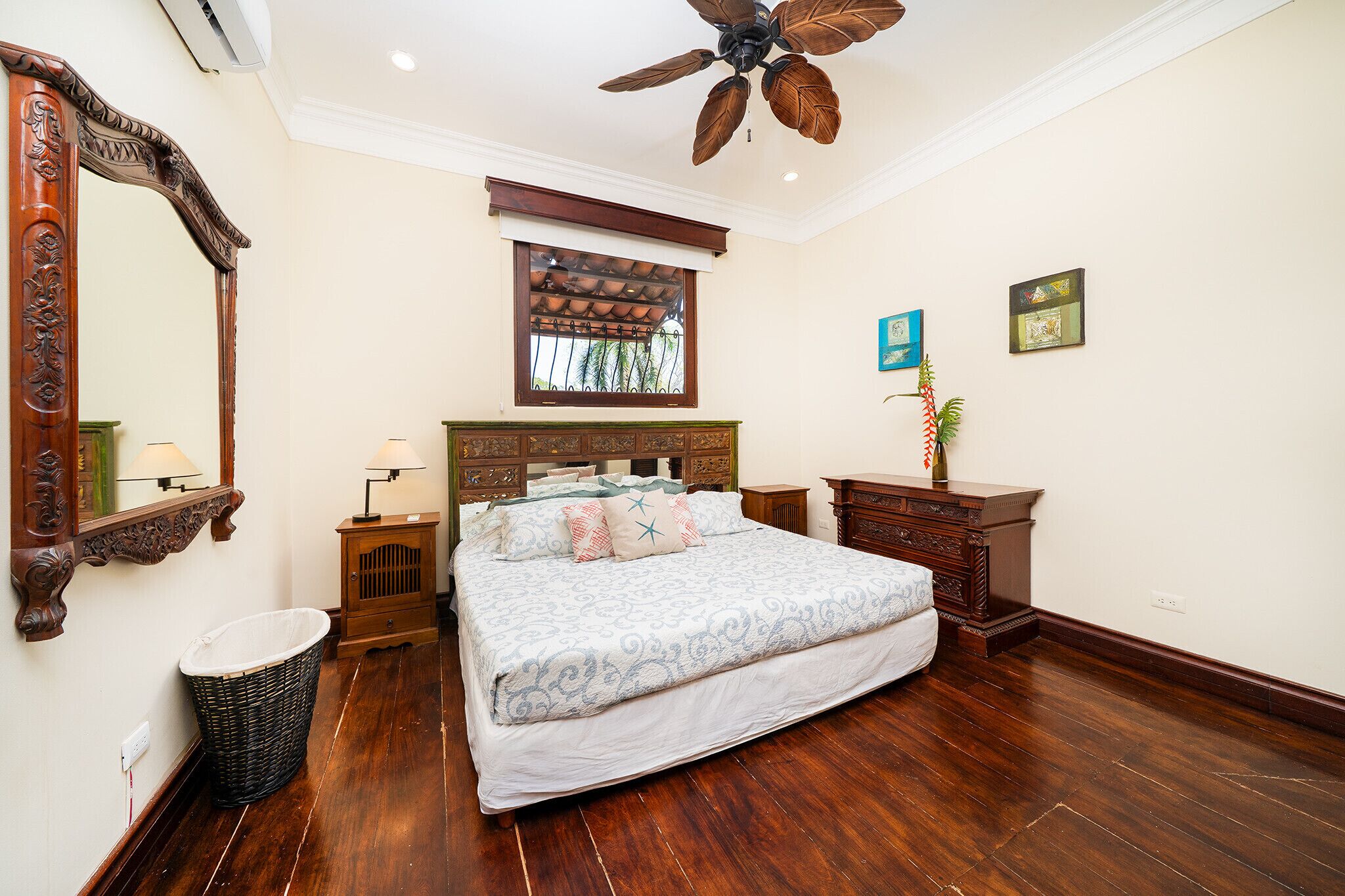 Une chambre avec un grand lit, une tête de lit en bois, un ventilateur de plafond, une fenêtre avec vue et un miroir fixé au mur.