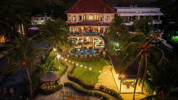 A large, two-story house with a swimming pool, surrounded by palm trees and a well-lit garden.
