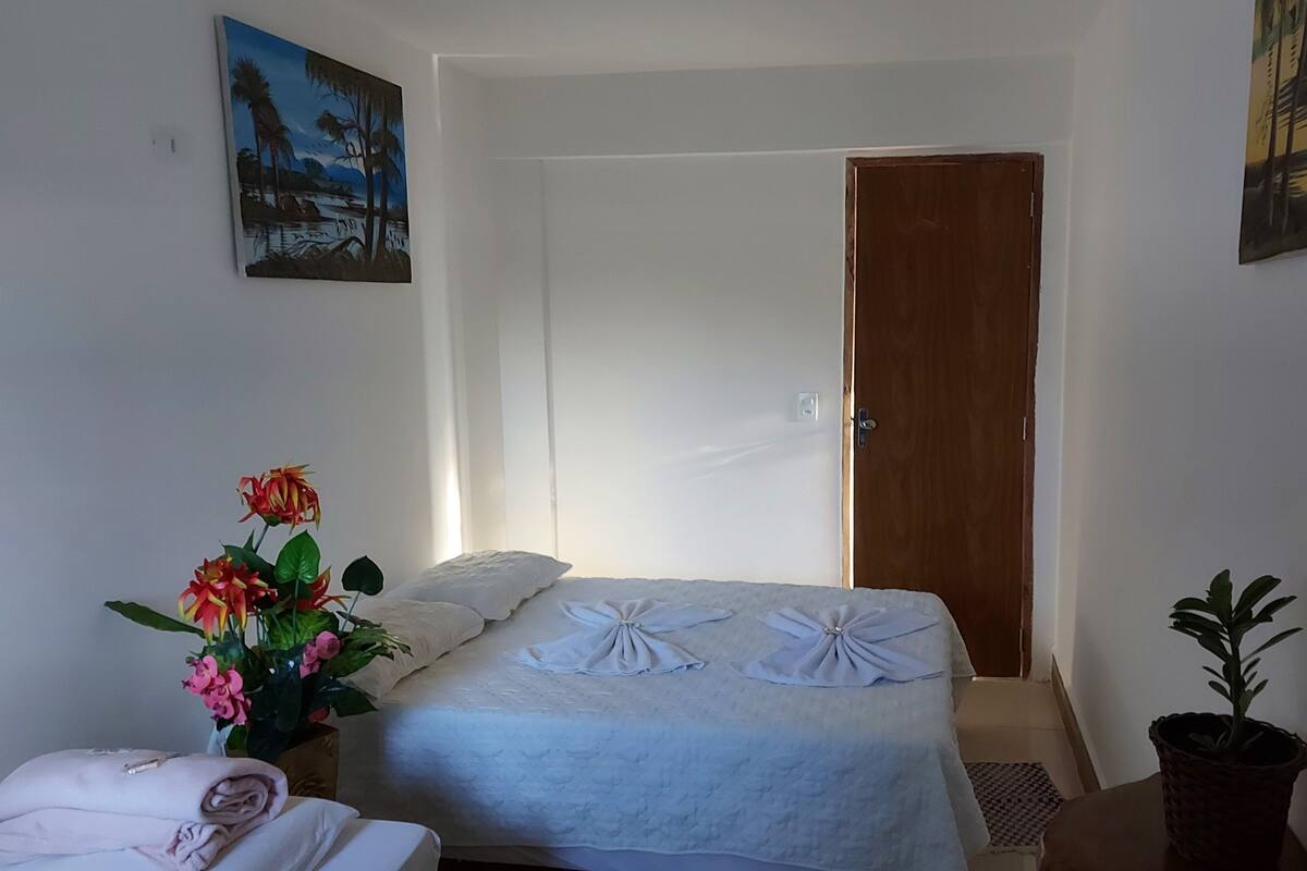 Une chambre à coucher avec un lit, une porte en bois, un tableau accroché au mur, une plante en pot et un vase rempli de fleurs.