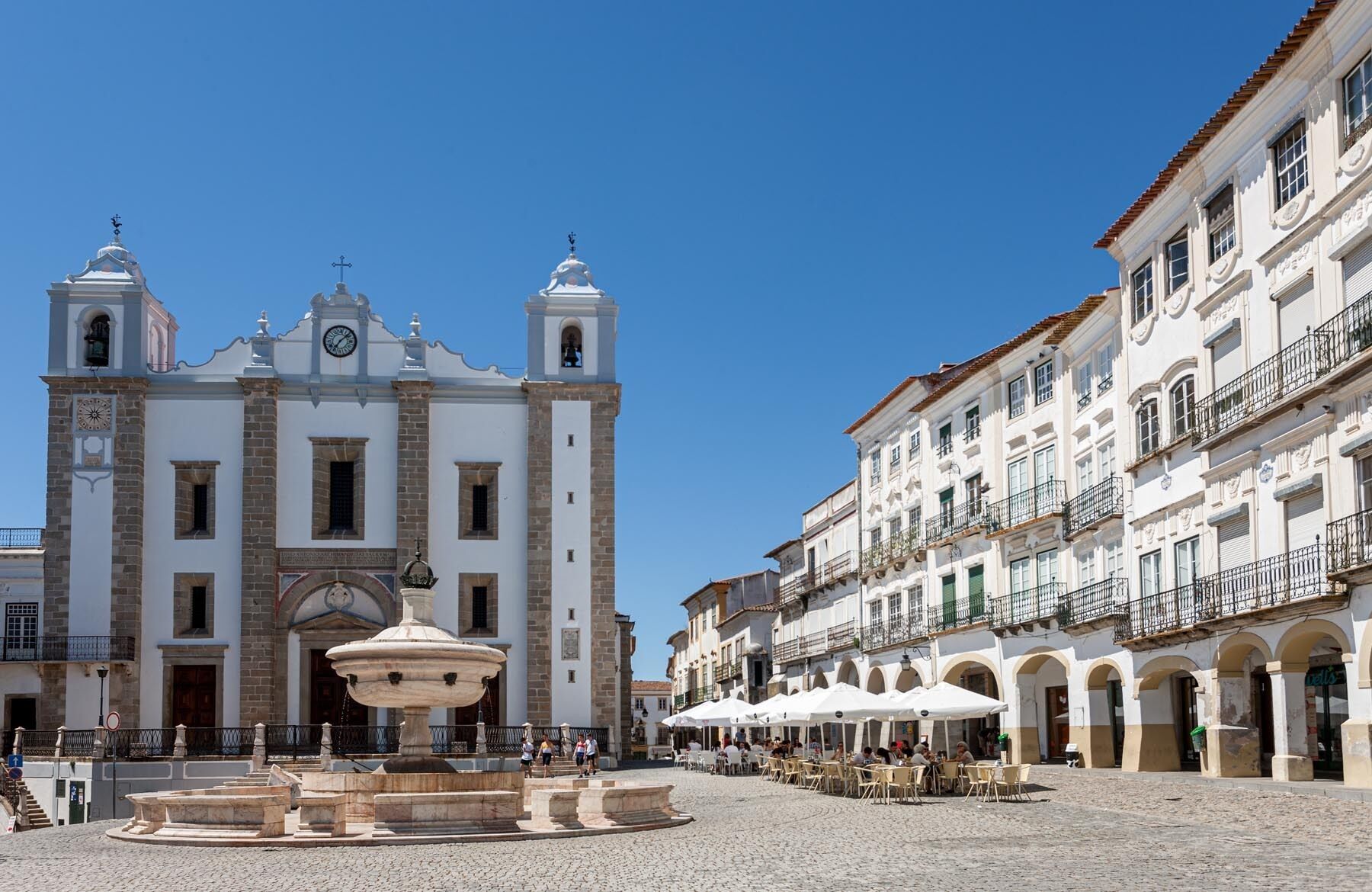 Une place historique abritant une église remarquable, avec une fontaine centrale, et entourée de bâtiments traditionnels dotés d’espaces extérieurs aménagés en terrasses.